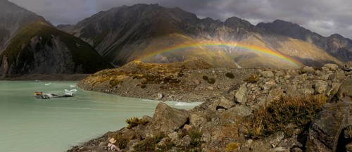 A vibrant rainbow arcs over Mount Cook in New Zealand during autumn. The scene captures the rugged