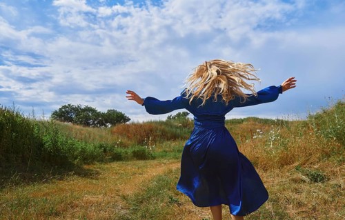 A joyful blond woman in a vibrant blue dress playfully twirls in a lush grassy field under a broad