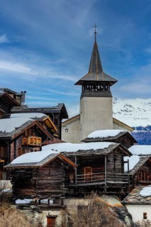 Old wooden houses with snow-covered roofs and the Saint Theodule church in the historic village