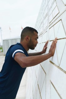 Black male athlete focuses intensely during an outdoor workout, pressing against a patterned wall.