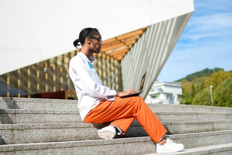A man in bright orange pants and a white shirt sits on outdoor stairs, focused on his laptop. The