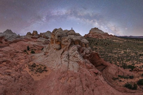 A distant, unrecognizable hiker stands atop a rugged rock formation under a star-filled sky in