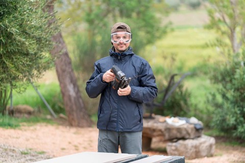 An happy man stands outdoors near a wooded area, holding a jigsaw for a carpentry project. He