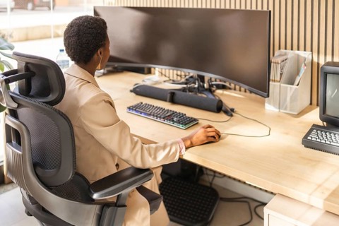 A person working at a modern office desk equipped with a large curved monitor, ergonomic chair, and