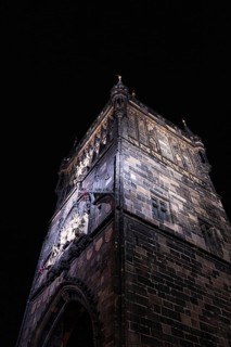 A dramatic view of a Gothic tower in Prague, captured at night. The architectural details are