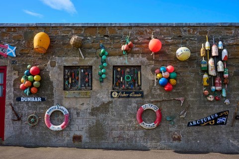 A stone wall decorated with colorful buoys and maritime elements against a blue sky, Dunbar, East