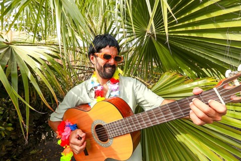 A cheerful man wearing sunglasses, a wig and a colorful flower necklace plays a guitar surrounded