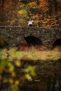 A woman stands on a stone bridge overlooking a tranquil pond surrounded by vivid autumn foliage in