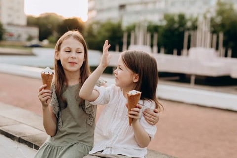 Two young girls joyfully share ice cream cones while sitting on a bench in a park, enjoying a sunny