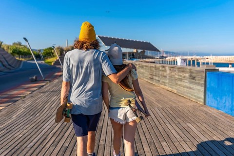 Young couple walking hand in hand on a sunny boardwalk, carrying skateboards and enjoying a