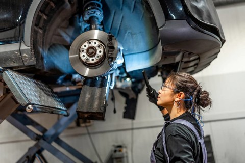 Female mechanic using flashlight, inspecting brakes of lifted car in a professional workshop,