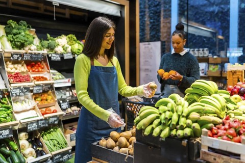 Supermarket employee wearing gloves arranging fresh kiwi fruit on display while customer chooses