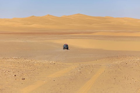 A car drives alone in a vast desert landscape with sand-coloured dunes and a clear sky, Off-road