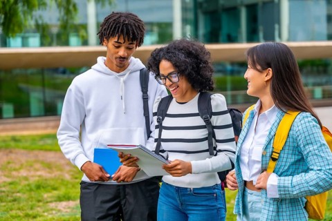 Three multiracial students smiling while reading notes standing outside the university