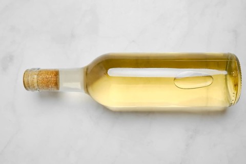 A glass bottle with white wine on a smooth light surface. The bottle has a cork top and shows the