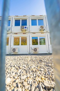 White containers with air conditioners on gravel surface, seen through a barrier system, in natural