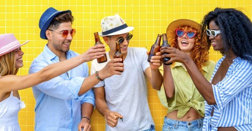 Five cheerful young friends wearing summer clothes and hats are toasting with beer bottles,