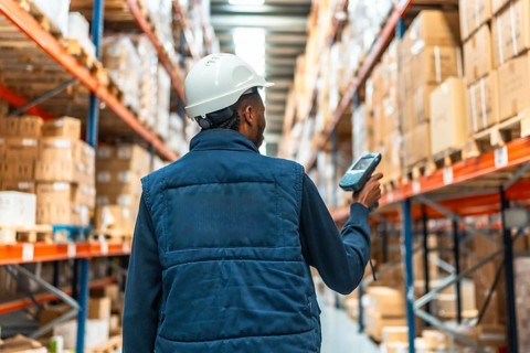 Rear view of an african worker with waistcoat and helmet scanning packages with wireless scanner in