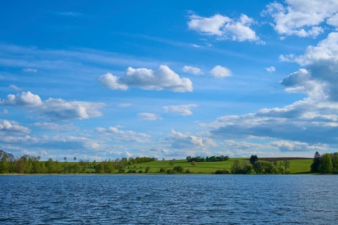 Ober Moosersee surrounded by green fields and trees under a partly cloudy blue sky, spring,