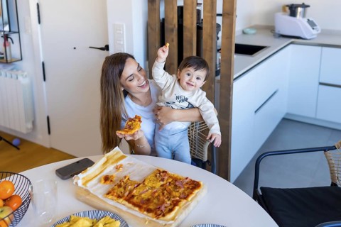 Happy mother holding her baby while eating pizza in a bright, modern kitchen, creating a