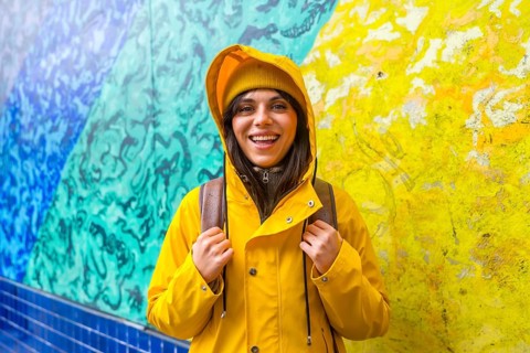 Young woman in a yellow raincoat and knit hat smiling brightly against a vibrant blue and yellow