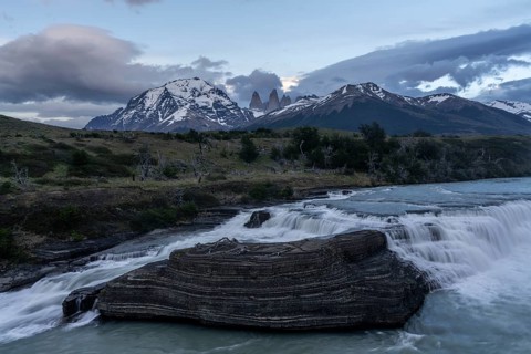 Cascades of the Río Paine with layered rocks in the foreground, behind them the snow-covered