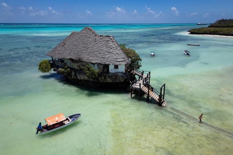 Aerial view of the building surrounded by turquoise water and white sand beach with boats under a