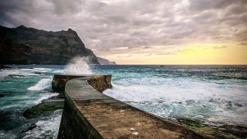 A pier leads into the sea under dramatic clouds and waves at sunset, Ponta do Sol, Santo Antao,