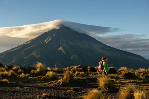 A woman in hiking gear stands before the majestic Mount Taranaki in New Zealand, basking in the