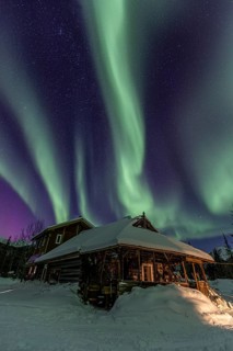 Green and purple northern lights (aurora borealis) over a log cabin in the snow, Brooks Range,