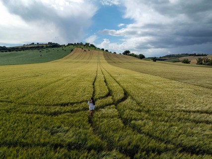 A young woman in a white dress walks through lush green fields in the Italian countryside. The