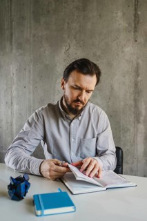 A man in a shirt sits at a table in a minimal office, writing in a notebook. A blue notebook and