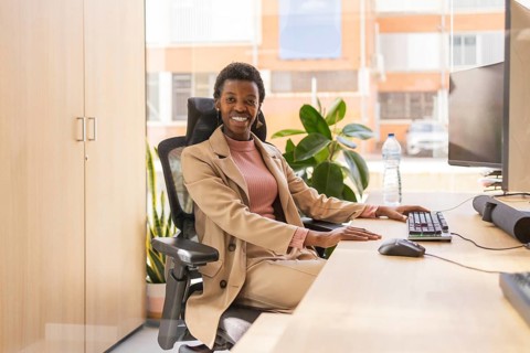 A person wearing a suit sits at a desk, smiling confidently. The office is modern with natural