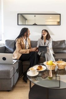 Two businesswomen are sitting on a sofa in a hotel room, laughing while working on a tablet during