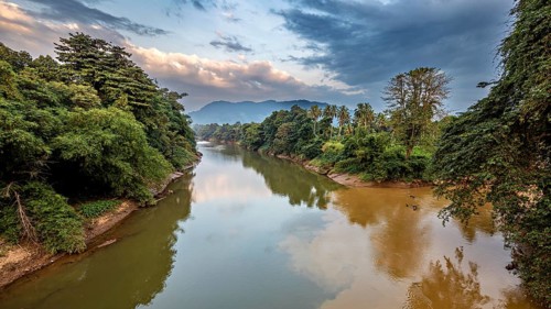 A river surrounded by lush vegetation and mountains in the background, including a cloudy sky, The