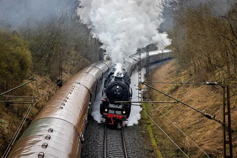 A steam locomotive with lots of smoke goes through a forest over winding tracks, historic steam
