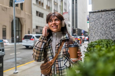 Woman holding a takeaway coffee cup and wearing headphones, engaging with her smartphone while