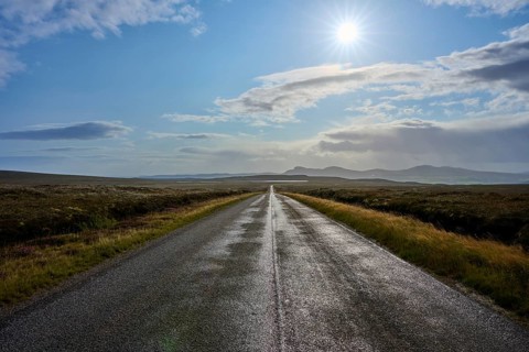 Long road under light blue sky with sun surrounded by beautiful countryside and mountains in the