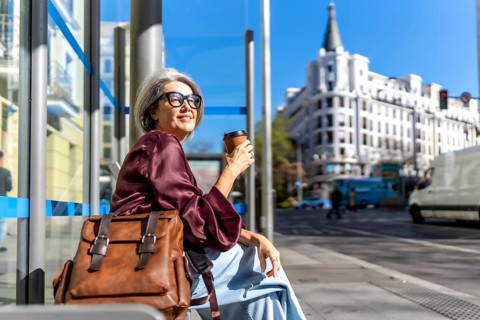 Senior woman is sitting at a city bus stop, holding a disposable coffee cup, embracing a relaxed