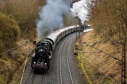 A steaming locomotive pulls a train through a winding wooded landscape, historic steam locomotive