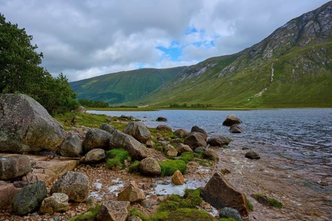Rocks on the shore of a lake with green mountains in the background under cloudy sky, Loch Etive,