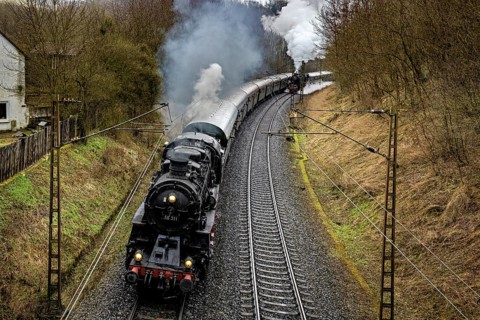 A black locomotive with steaming smoke follows winding tracks through nature, historic steam