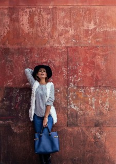 Stylish woman wearing a hat and white vest poses with a large blue handbag against a textured,