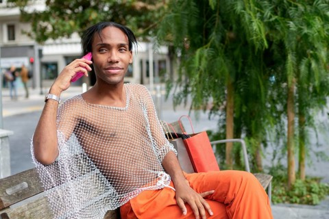 A stylish man sits on a bench, conversing on a pink smartphone. His fashion-forward mesh shirt and