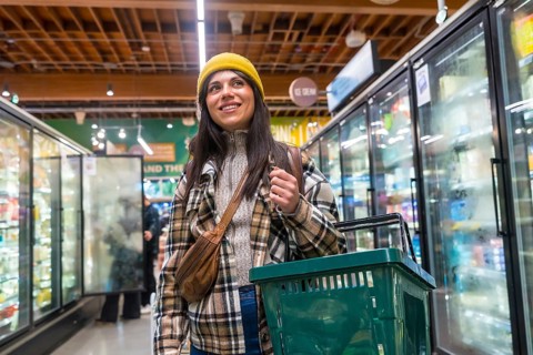 Young woman wearing a yellow beanie and plaid jacket smiling and carrying a shopping basket while
