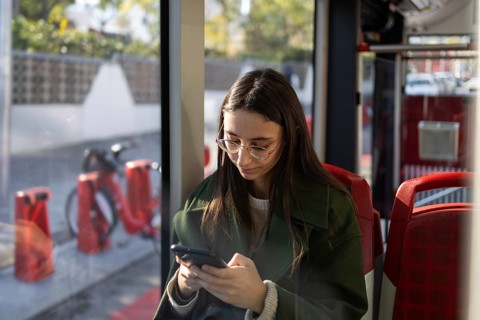 A young woman wearing glasses and a green coat looks at her smartphone while riding public