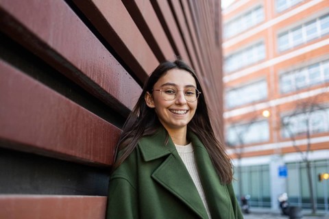 Smiling woman in a green coat leans against a red wall in an urban setting Her relaxed expression