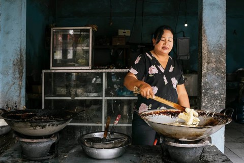 A woman prepares traditional street food in a market setting of Bali. She is frying snacks in a