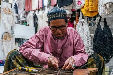 A balinese artisan wearing glasses focuses on crafting handmade goods in a vibrant local market.