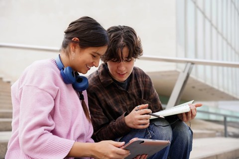 Two teenagers, a non-binary individual and a female, are studying together outdoors, using a tablet